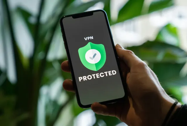 Una mano sostiene un celular cuya pantalla muestra un ícono de escudo verde con un tilde y el texto 'VPN Protected'